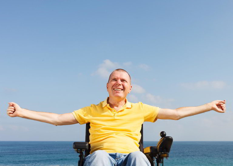 Man in wheelchair enjoying life, smiling with arms extended, sitting at the beach in from of the ocean.